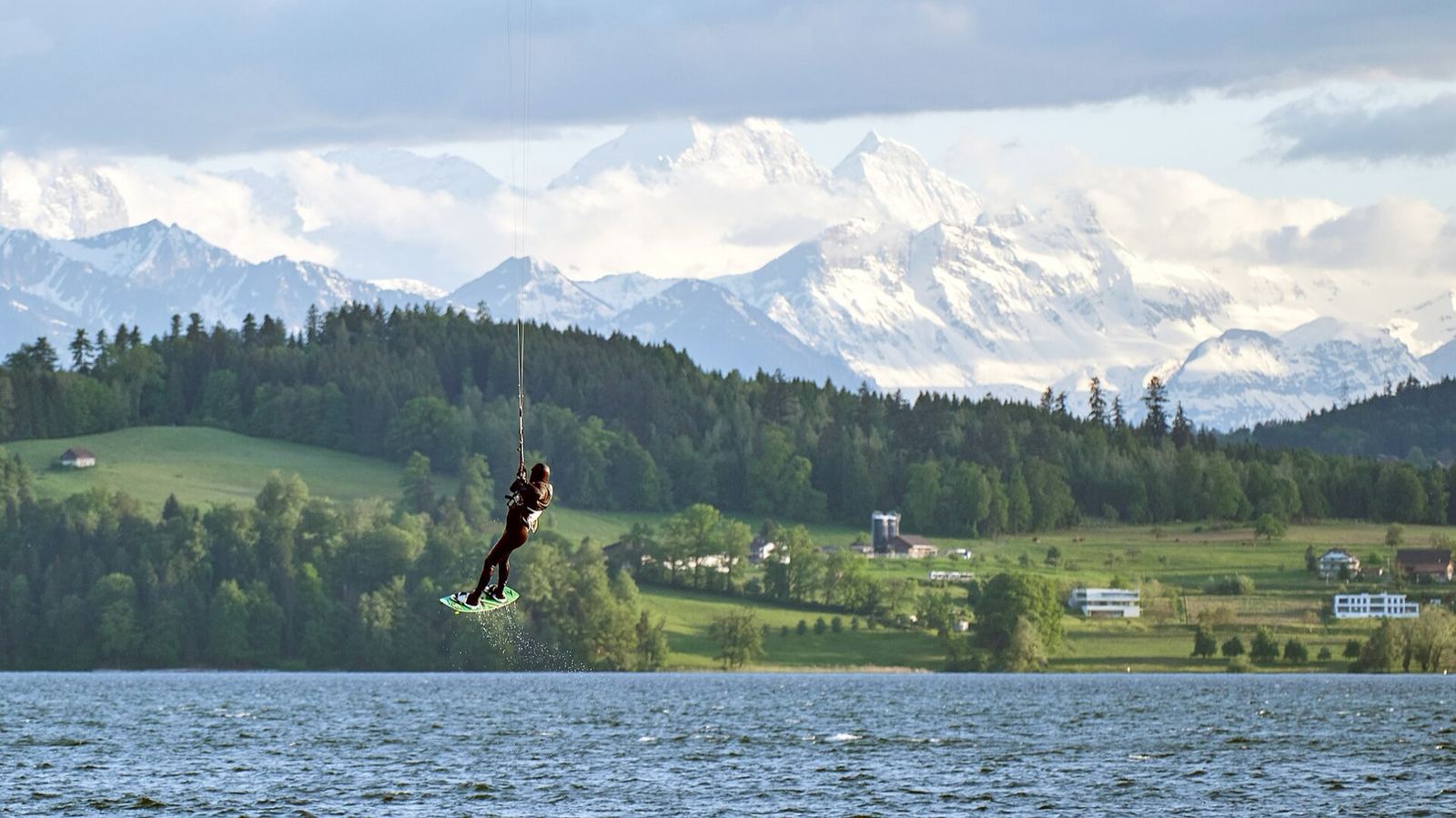 Kitesurfista saltando sobre un lago con montañas nevadas de fondo